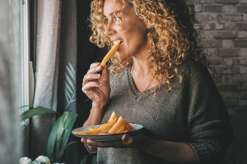 Beautiful adult woman with curly hair stands at home eating melon slices, smiling dreamily